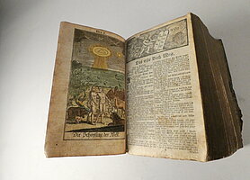Die Bibel ist aufgeschlagen. Links ist ein Bild. Es zeigt Adam und Eva und die Schöpfung der Welt. Rechts beginnt das erste Kapitel der Bibel.
