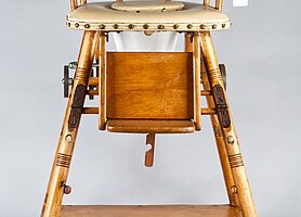 Der Stuhl ist aus Holz. Er ist hoch. Die Sitzfläche ist gepolstert. Sie hat ein Loch, so kann der Stuhl auch eine Toilette sein. Vorne ist eine Klappe, auf der die Füße abgestellt werden können. Der Stuhl kann zu einem kleineren Stuhl mit Tisch umgebaut werden.