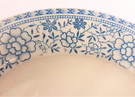 Der Teller hat einen breiten Rand. Der Rand ist mit einem blauen Muster verziert. Das Muster zeigt Blüten und Blätter.