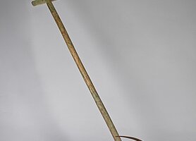 Das Gerät hat einen Holzstiel. Am Stiel ist eine Schippe. Die Schippe hat einen Metallbogen. Unten an der Schippe sind Kufen.