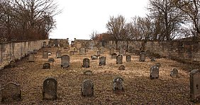 Auf dem Friedhof stehen viele Grabsteine. Die meistens sind oben rund. Um den Friedhof ist eine Mauer.
