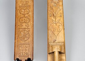 Es sind zwei Bretter. Sie haben Griffe. Beide Bretter sind verziert. Sie zeigen Muster, Sterne, Herzen, Blumen, Jahreszahlen und Buchstaben.